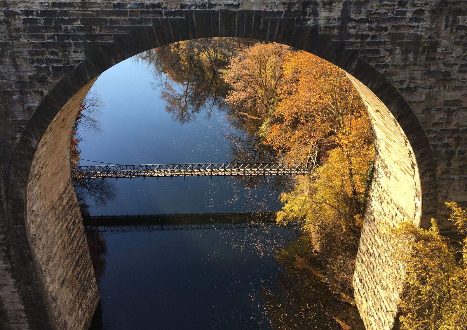 Swinging Bridge over the Brandywine River framed by a stone railroad arch, with orange and gold fall trees
