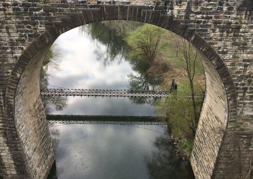 Swinging Bridge through a stone railroad arch in spring, pale green trees and clouds reflected in the river