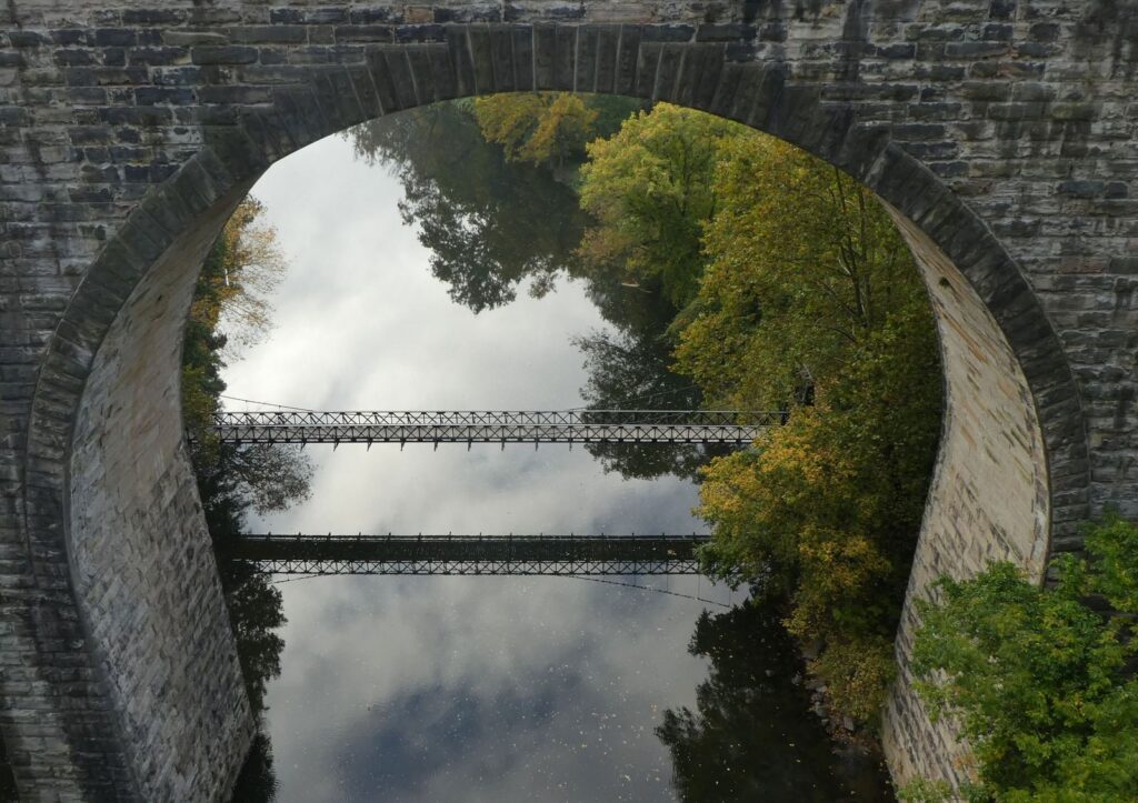 Swinging Bridge through a stone railroad arch in summer, full green trees and clouds reflected in the river