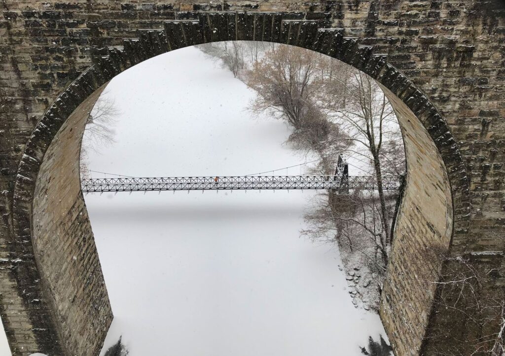 Swinging Bridge framed by a stone railroad arch over a snow-covered Brandywine River, bare trees along the bank